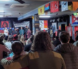 Audience at Semaphore Workers Club event, with speakers on stage and flags overhead.