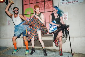 Three people celebrate in SWARM In The Streets with tutus and kilts near a "no smoking" sign.
