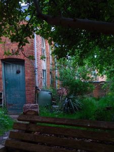 Blackcurrent Centre garden view with brick building, green door, rain barrel, and bench.